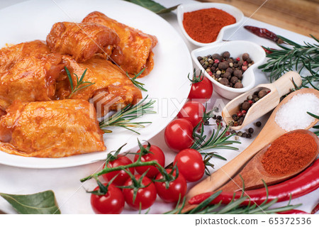 Chicken legs in a red marinade on a white plate with wooden spoons with seasonings for cooking, a branch of razmarin, a branch of red tomatoes. Chicken legs in a red marinade on a white plate with wooden spoons with seasonings for cooking, a branch of razmarin, a branch of red tomatoes. 65372536