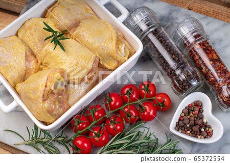 Chicken legs in a yellow marinade in a white baking plate with a branch of razmarin, spices, hot pepper and a branch of tomatoes on a marble kitchen board.Raw Chicken thigh before cooking. Chicken legs in a yellow marinade in a white baking plate with a branch of razmarin, spices, hot pepper and a branch of tomatoes on a marble kitchen board.Raw Chicken thigh before cooking. 65372554