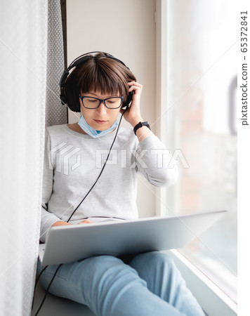 Woman in medical mask remote works from home. She sits on window sill with laptop on knees and headphones. Lockdown quarantine because of coronavirus COVID19. Self isolation at home. Woman in medical mask remote works from home. She sits on window sill with laptop on knees and headphones. Lockdown quarantine because of coronavirus COVID19. Self isolation at home. 65372841