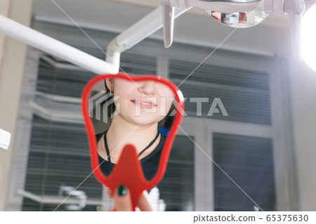 Happy female patient looking at mirror in stomatology clinic. Dentistry, dental office and medicine concept. 65375630