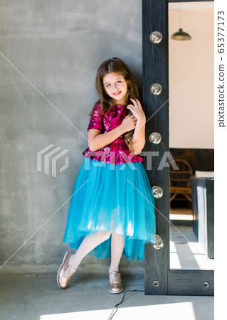 Beautiful cute little girl in a pink shiny blouse and a blue skirt on a grey background standing near mirror. Beautiful cute little girl in a pink shiny blouse and a blue skirt on a grey background standing near mirror. 65377173