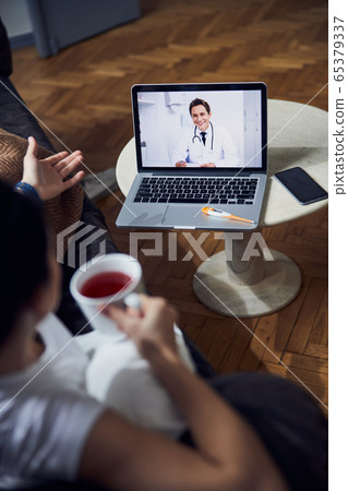 Caucasian ill woman with cup in arm listening to doctor on laptop Caucasian ill woman with cup in arm listening to doctor on laptop 65379337