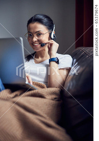 Caucasian smiling lady in headphones sitting with laptop on coach indoors 65379674