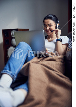 Caucasian positive lady in headphones sitting on coach with laptop Caucasian positive lady in headphones sitting on coach with laptop 65379676