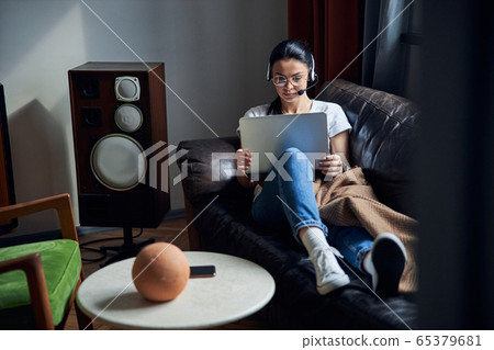 Caucasian serious lady in headphones sitting on sofa with laptop Caucasian serious lady in headphones sitting on sofa with laptop 65379681