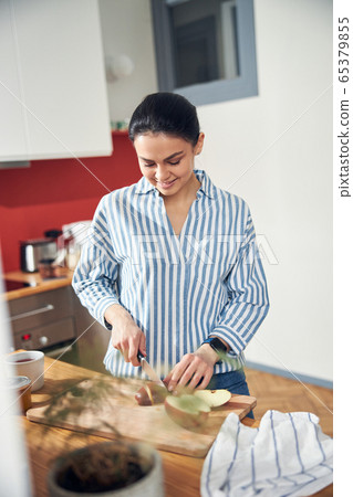 Beautiful Caucasian lady in striped shirt cutting apple into pieces in kitchen Beautiful Caucasian lady in striped shirt cutting apple into pieces in kitchen 65379855