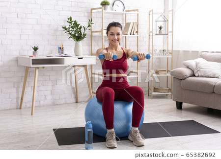 Strength training at home. Millennial girl exercising with dumbbells and fitness ball in living room 65382692