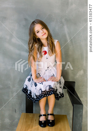 Beautiful little girl with long broun hair and blue eyes sitting on the chair on a grey background wearing a black and white spring dress 65383477
