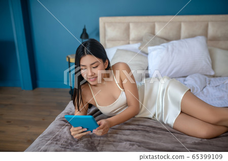 Young brunette enjoying her morning, lying on her side, checking something on her tablet, smiling Young brunette enjoying her morning, lying on her side, checking something on her tablet, smiling 65399109
