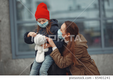 A European mother in a respirator with her daughter are standing near a building.The parent is teaching her child how to wear protective mask to save herself from virus 65401241