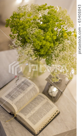 Composition: an open Bible, a bouquet of wildflowers in a vase and a decorative flashlight with a candle on the nightstand against the background of burning lights. Composition: an open Bible, a bouquet of wildflowers in a vase and a decorative flashlight with a candle on the nightstand against the background of burning lights. 65408843