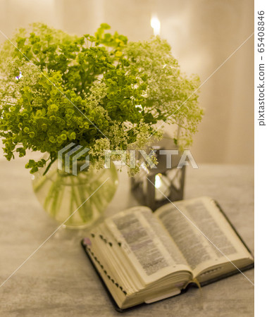 Composition: an open Bible, a bouquet of wildflowers in a vase and a decorative flashlight with a candle on the nightstand against the background of burning lights. 65408846