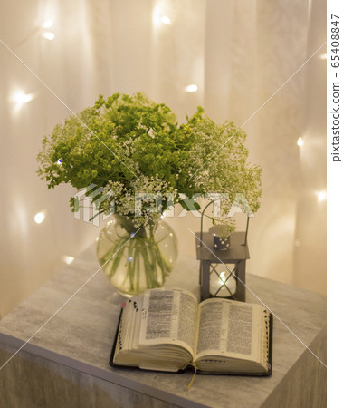 Composition: an open Bible, a bouquet of wildflowers in a vase and a decorative flashlight with a candle on the nightstand against the background of burning lights. Composition: an open Bible, a bouquet of wildflowers in a vase and a decorative flashlight with a candle on the nightstand against the background of burning lights. 65408847