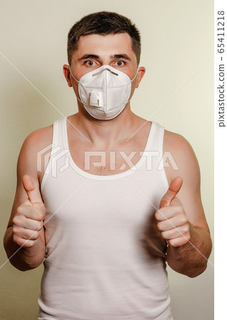 Patient man in medical mask on self isolation during quarantine and coronavirus pandemic stands with arms crossed. Shows a thumbs-up of both hands. 65411218