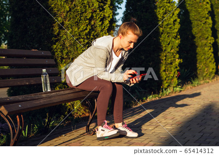 Young female runner, athlete resting after jogging in the city street in sunshine. Beautiful caucasian woman training, listening to music Young female runner, athlete resting after jogging in the city street in sunshine. Beautiful caucasian woman training, listening to music 65414312