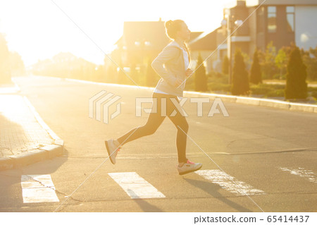 Young female runner, athlete is jogging in the city street in sunshine. Beautiful caucasian woman training, listening to music 65414437