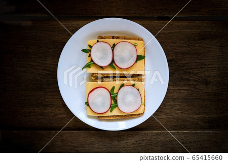 Sandwich made from rye bread, hard cheese, radish, arugula, served on a white plate against a dark 65415660