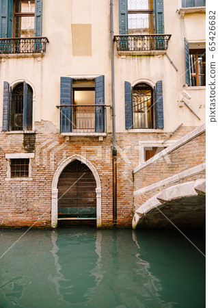 A brick building in the water in Italy, Venice. A brick bridge over a small narrow canal, classic Venetian windows in the facade of the building with wooden shutters and forged bars. 65426682