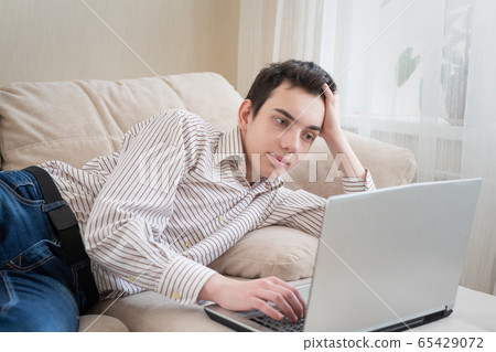 Young man lying on sofat and using laptop at home 65429072