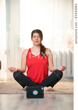 Woman practices yoga and meditates in the lotus position at home using video tutorials. 65440864