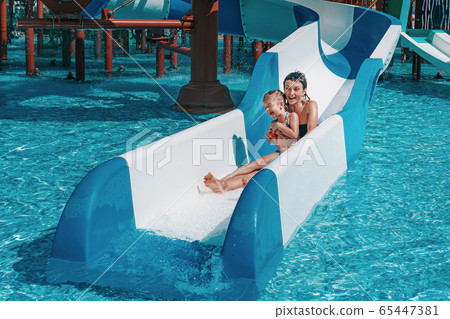 Little girl in swimsuit goes down the blue slides to the pool, Mom and daughter play and swim in the outdoor pool 65447381