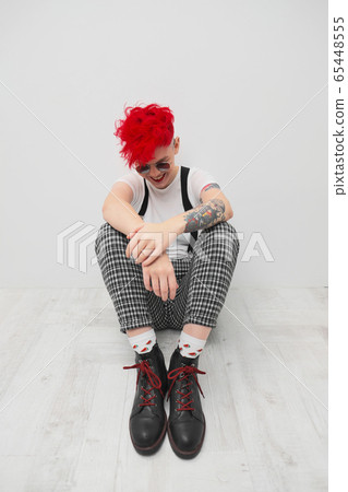 Portrait of a cool fashionable modern young girl. A short haircut with shaved temple. Dyed bright red hair. Red lipstick. Studio photo on white background. Suspenders and checkered pants. Portrait of a cool fashionable modern young girl. A short haircut with shaved temple. Dyed bright red hair. Red lipstick. Studio photo on white background. Suspenders and checkered pants. 65448555