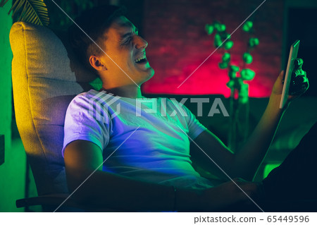 Cinematic portrait of handsome young man using devices, gadgets in neon lighted interior. Youth culture, bright colors Cinematic portrait of handsome young man using devices, gadgets in neon lighted interior. Youth culture, bright colors 65449596