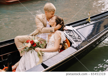 Italy wedding in Venice. The bride and groom ride in a classic wooden gondola along a narrow Venetian canal. Close-up of cuddles newlyweds. Italy wedding in Venice. The bride and groom ride in a classic wooden gondola along a narrow Venetian canal. Close-up of cuddles newlyweds. 65451319