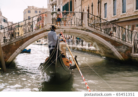 Italy wedding in Venice. A gondolier rolls a bride and groom in a classic wooden gondola along a narrow Venetian canal. Gondola swims under an arched bridge, rear view. Italy wedding in Venice. A gondolier rolls a bride and groom in a classic wooden gondola along a narrow Venetian canal. Gondola swims under an arched bridge, rear view. 65451351