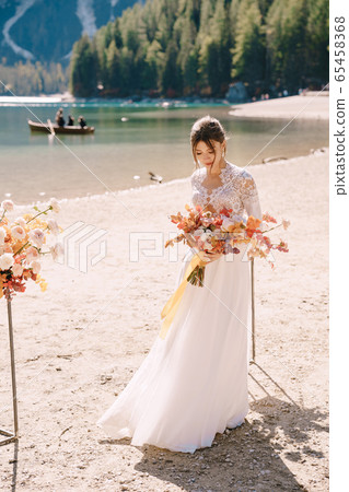 Beautiful bride in a white dress with sleeves and lace, with a yellow autumn bouquet on background of the arch for ceremony, at Lago di Braies in Italy. Destination wedding in Europe, on Braies lake. Beautiful bride in a white dress with sleeves and lace, with a yellow autumn bouquet on background of the arch for ceremony, at Lago di Braies in Italy. Destination wedding in Europe, on Braies lake. 65458368