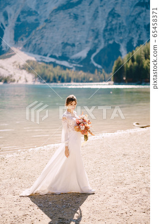 Beautiful bride in a white dress with sleeves and lace, with a yellow autumn bouquet of dried flowers and peony roses, on the Lago di Braies in Italy. Destination wedding in Europe, on Braies lake. Beautiful bride in a white dress with sleeves and lace, with a yellow autumn bouquet of dried flowers and peony roses, on the Lago di Braies in Italy. Destination wedding in Europe, on Braies lake. 65458371