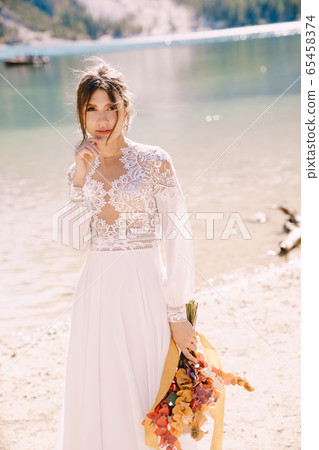 Beautiful bride in a white dress with sleeves and lace, with a yellow autumn bouquet of dried flowers and peony roses, on the Lago di Braies in Italy. Destination wedding in Europe, on Braies lake. Beautiful bride in a white dress with sleeves and lace, with a yellow autumn bouquet of dried flowers and peony roses, on the Lago di Braies in Italy. Destination wedding in Europe, on Braies lake. 65458374