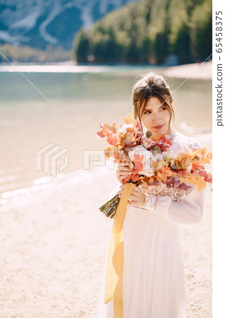 Beautiful bride in a white dress with sleeves and lace, with a yellow autumn bouquet of dried flowers and peony roses, on the Lago di Braies in Italy. Destination wedding in Europe, on Braies lake. 65458375