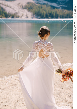Beautiful bride in a white dress with sleeves and lace, with a yellow autumn bouquet of dried flowers and peony roses, on the Lago di Braies in Italy. Destination wedding in Europe, on Braies lake. Beautiful bride in a white dress with sleeves and lace, with a yellow autumn bouquet of dried flowers and peony roses, on the Lago di Braies in Italy. Destination wedding in Europe, on Braies lake. 65458376