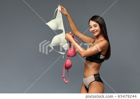 A difficult choice, a beautiful girl chooses a bra, holding bras of different colors in her hands. Gray background, copy space, make difficult choices. A difficult choice, a beautiful girl chooses a bra, holding bras of different colors in her hands. Gray background, copy space, make difficult choices. 65479244