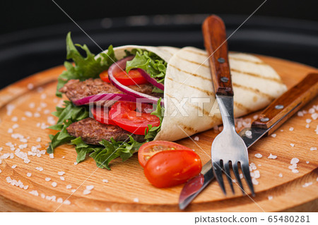 Beef roll, cooked on fire in a thin dough tortilla with vegetables and salad. Served serve with cutlery on a wooden cutting board and sauce. Beef roll, cooked on fire in a thin dough tortilla with vegetables and salad. Served serve with cutlery on a wooden cutting board and sauce. 65480281
