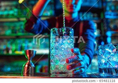 Close up of barman finishes preparation of alcoholic cocktail in multicolored neon light Close up of barman finishes preparation of alcoholic cocktail in multicolored neon light 65481118