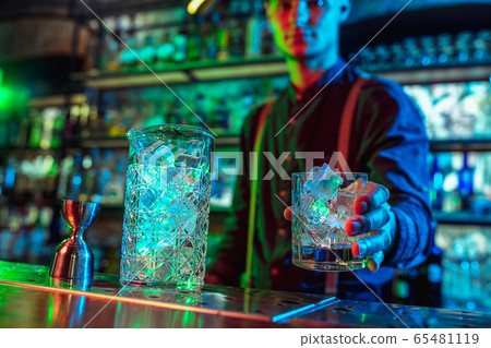 Close up of barman finishes preparation of alcoholic cocktail in multicolored neon light 65481119