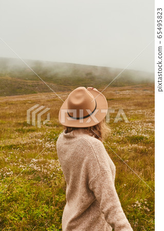 Enjoying wild nature. Rear view of a woman, female traveler in a hat and wool cardigan standing in mountain field on a cloudy, foggy da 65495823
