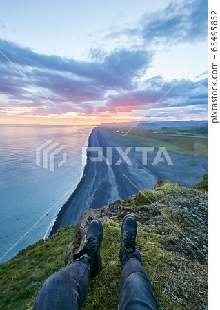 Traveler sitting on the cape above amazing black sand beach in Iceland and watching sunset or sunrise with soft pink clouds. Legs hanging from the cliff 65495852