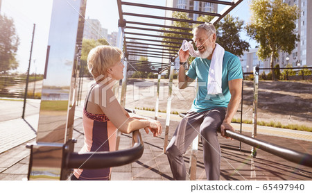 Tired mature couple resting after morning workout outdoors. They are standing near parallel bars and discussing something. Active senior couple doing sport together 65497940