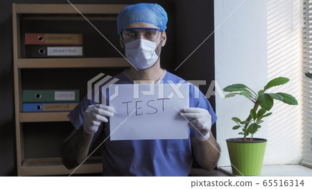 Doctor offers to make medical test. Man in blue protective uniform stands near window looking at camera while holding slogan test on paper sheet. Coronavirus pandemic concept Doctor offers to make medical test. Man in blue protective uniform stands near window looking at camera while holding slogan test on paper sheet. Coronavirus pandemic concept 65516314