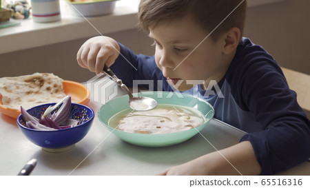 Hungry child learns to eat by himself. Little boy sits at dining table holding a spoon in his hands and eats soup. High angle view 65516316