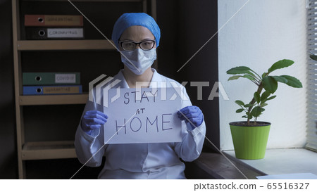 Stay at home slogan from tired doctor. Pandemic of Coronavirus concept. Female medic stands in medical office holding white paper blank 65516327