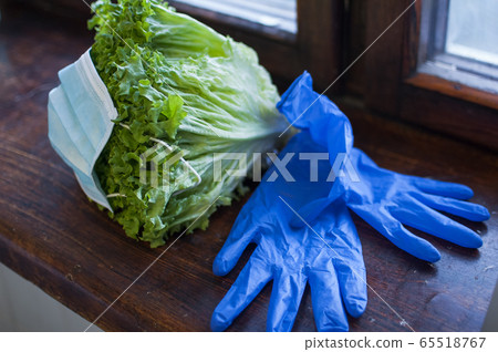 Green leaves of the salad in surgical mask lying on the wooden windowsill near blue protective gloves. Proper nutrition, delivery food during a Coronavirus pandemic and isolation. Green leaves of the salad in surgical mask lying on the wooden windowsill near blue protective gloves. Proper nutrition, delivery food during a Coronavirus pandemic and isolation. 65518767