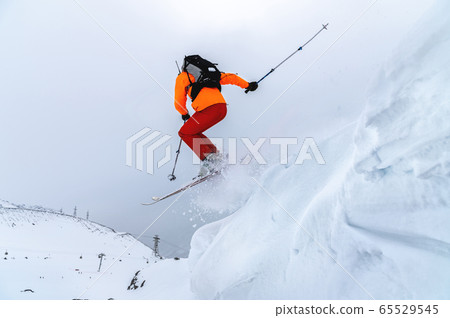 Male athlete skier in an orange trigger with a gray backpack and ski poles jumps from a high snowy slope against the backdrop of mountains in the clouds on a cloudy day. Male athlete skier in an orange trigger with a gray backpack and ski poles jumps from a high snowy slope against the backdrop of mountains in the clouds on a cloudy day. 65529545