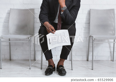 Closeup view of black guy in office suit with resume waiting for job interview at company foyer 65529906