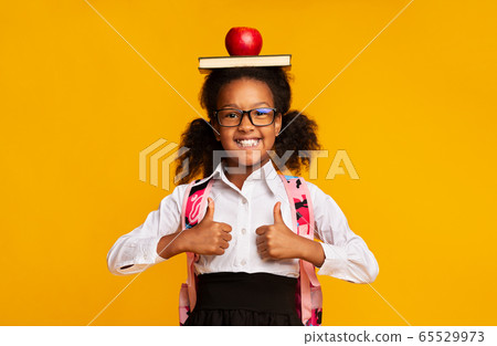 Schoolgirl Holding Book And Apple On Head Gesturing Thumbs-Up, Studio Schoolgirl Holding Book And Apple On Head Gesturing Thumbs-Up, Studio 65529973