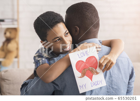 Little afro boy hugging dad and giving him handmade postcard Little afro boy hugging dad and giving him handmade postcard 65530201