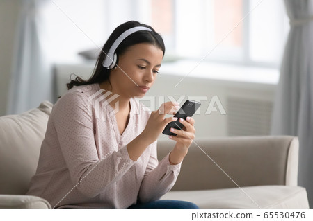 African woman sitting on couch wearing headphones using smartphone African woman sitting on couch wearing headphones using smartphone 65530476